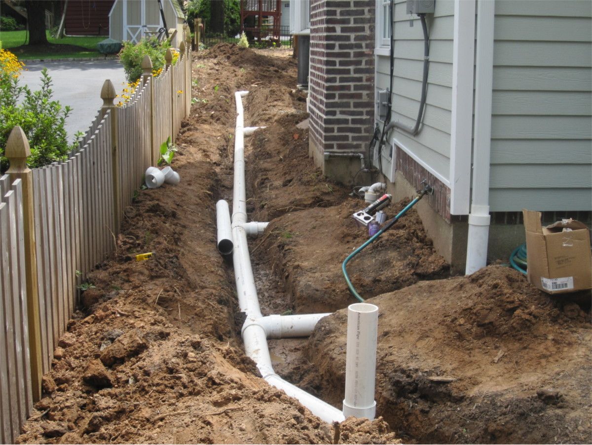 White drainage pipe laid in a narrow dirt trench beside a house and fence during construction work