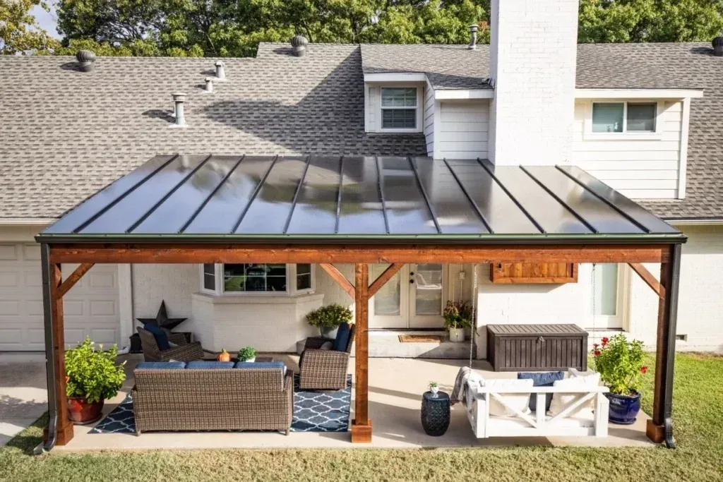Covered patio with black metal roof, wicker sofa, white benches, and potted plants behind a house