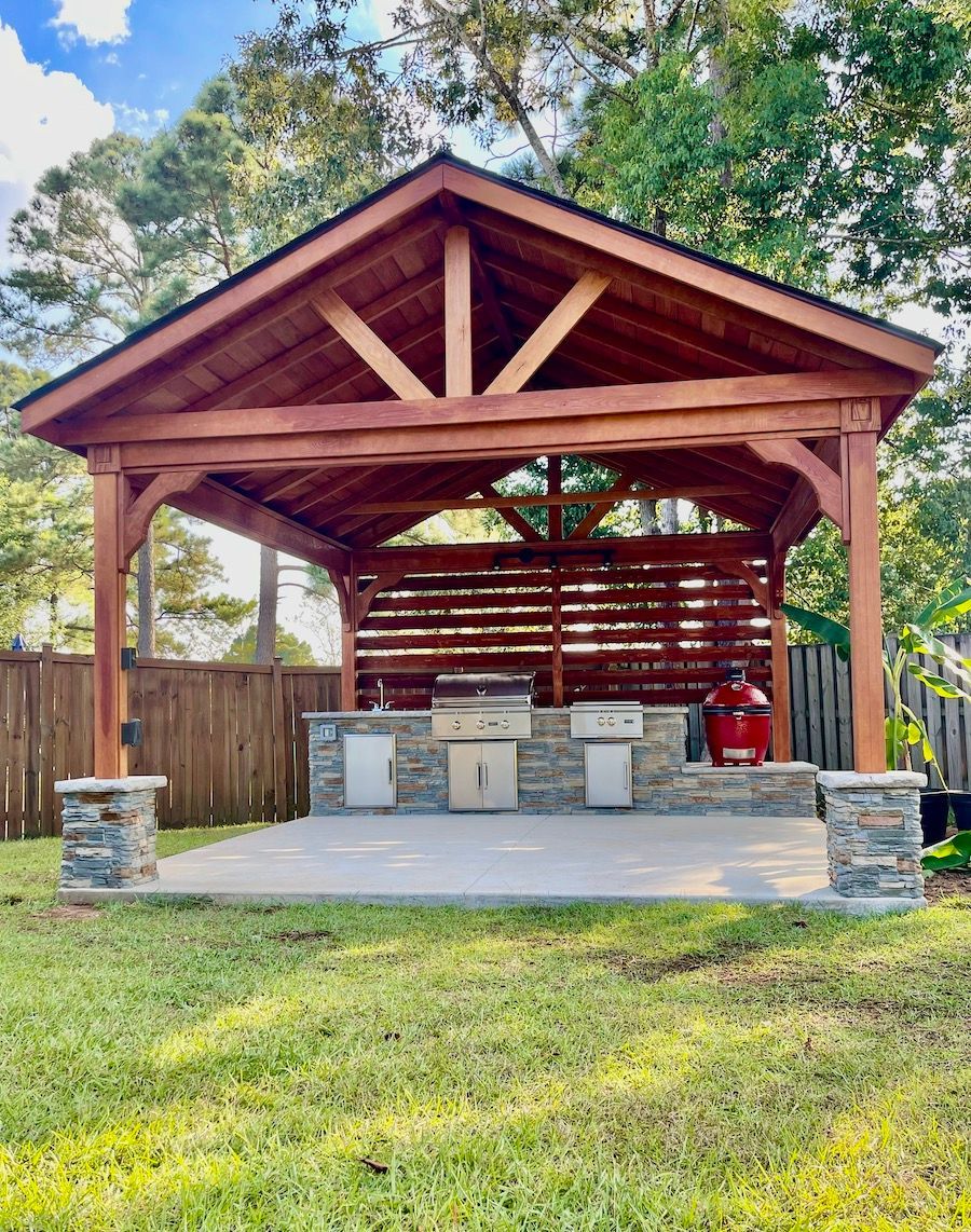 Open-air pavilion with wooden roof, stone columns, grills, and picnic tables in a grassy park.
