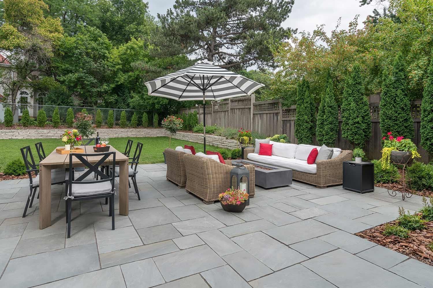 Patio with wicker seating, outdoor dining table, umbrella, and stone pavers in a landscaped backyard