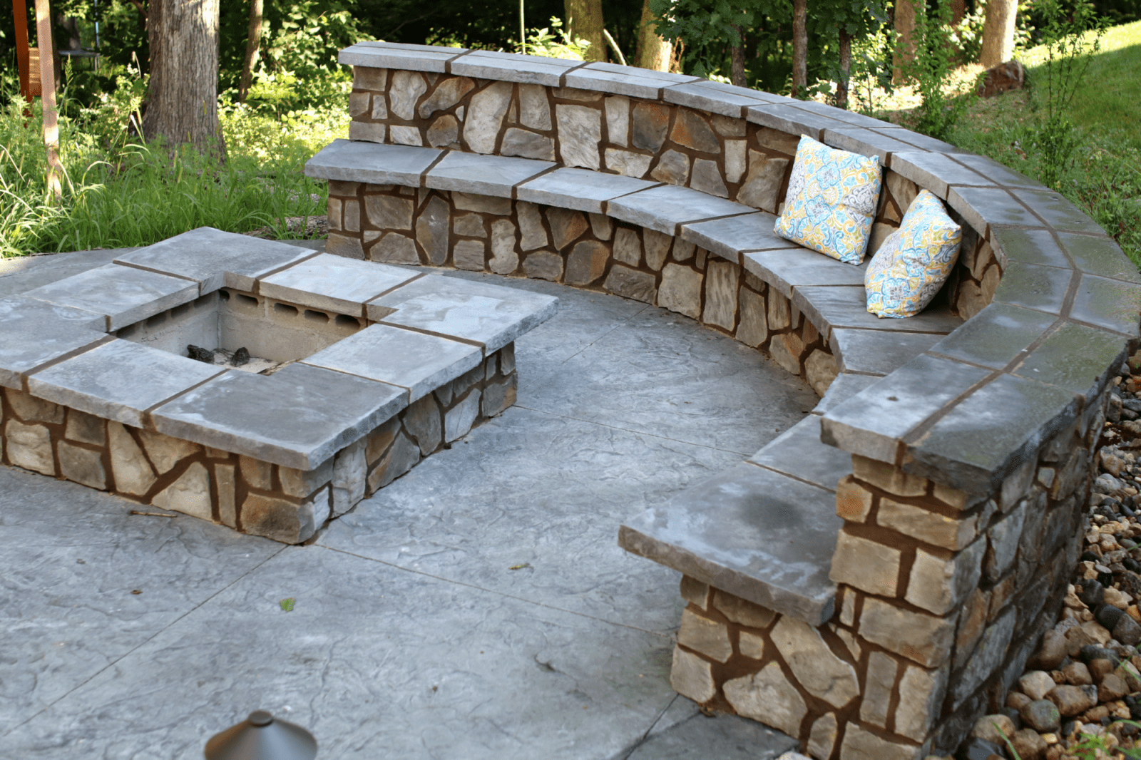 Stone outdoor fire pit with curved built-in bench seating and cushions in a wooded patio area