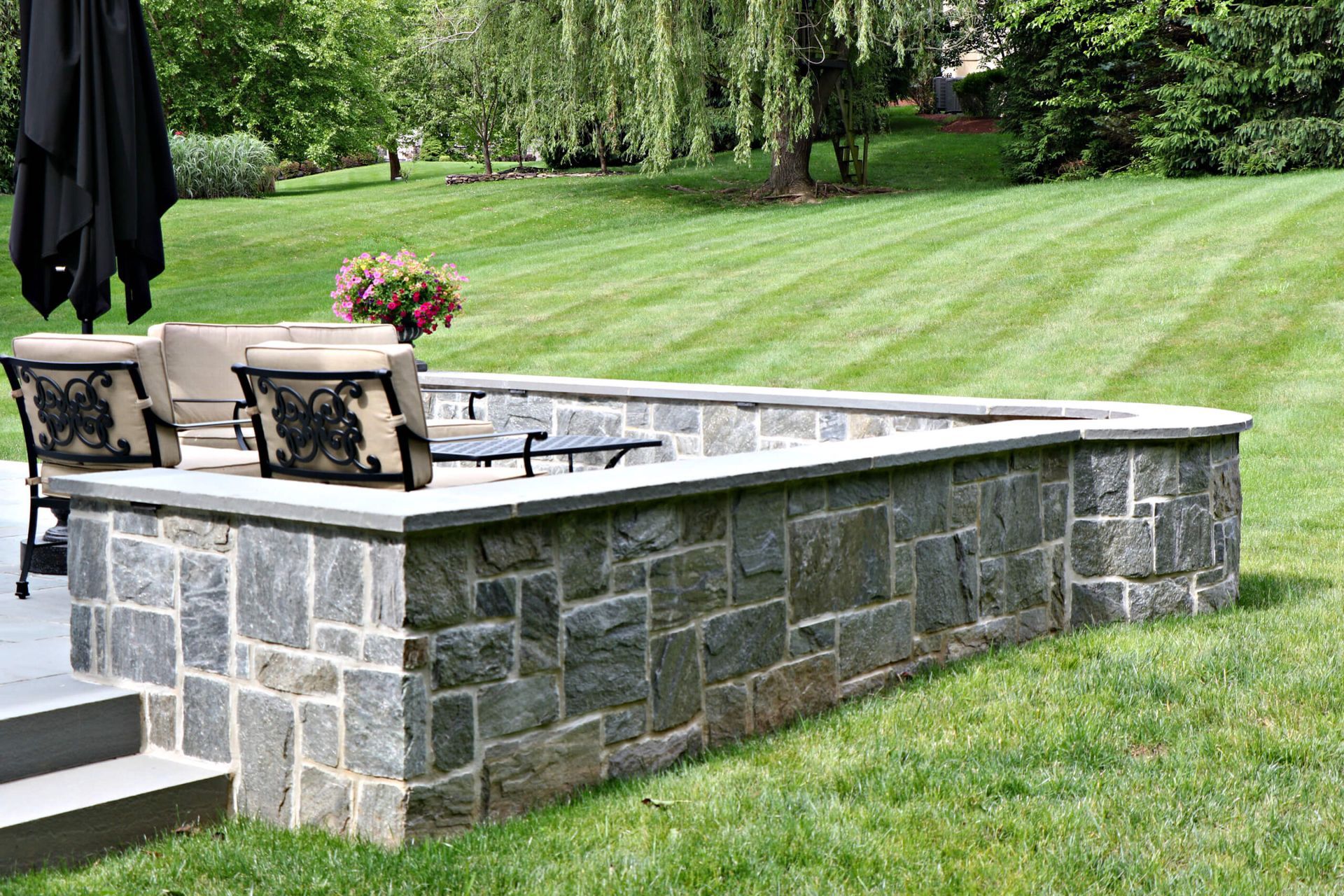 Stone patio seating area with two cushioned chairs and a raised stone wall overlooking a green lawn.