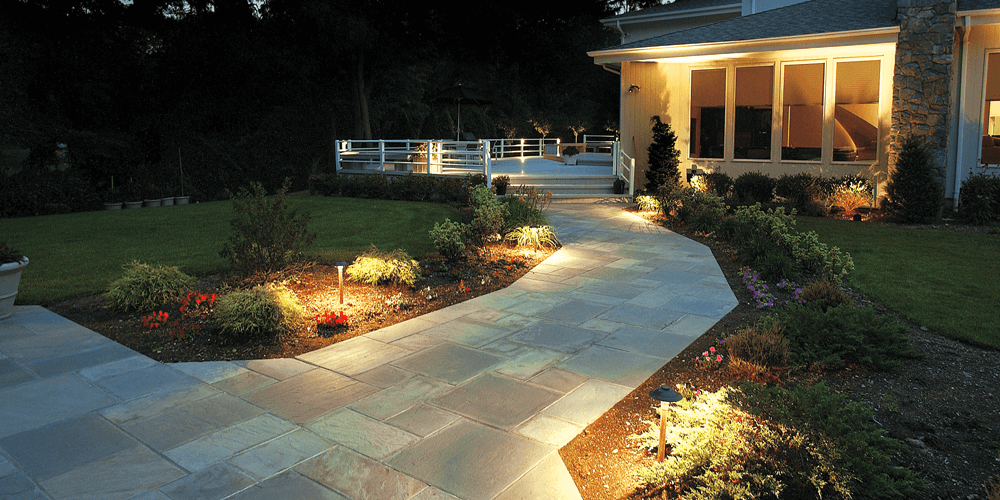 Curved stone walkway through a lit garden leading to a warmly lit house at night