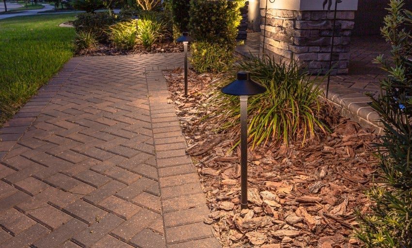 A lit garden path beside a house, with brick pavers, mulch, plants, and small landscape lights.