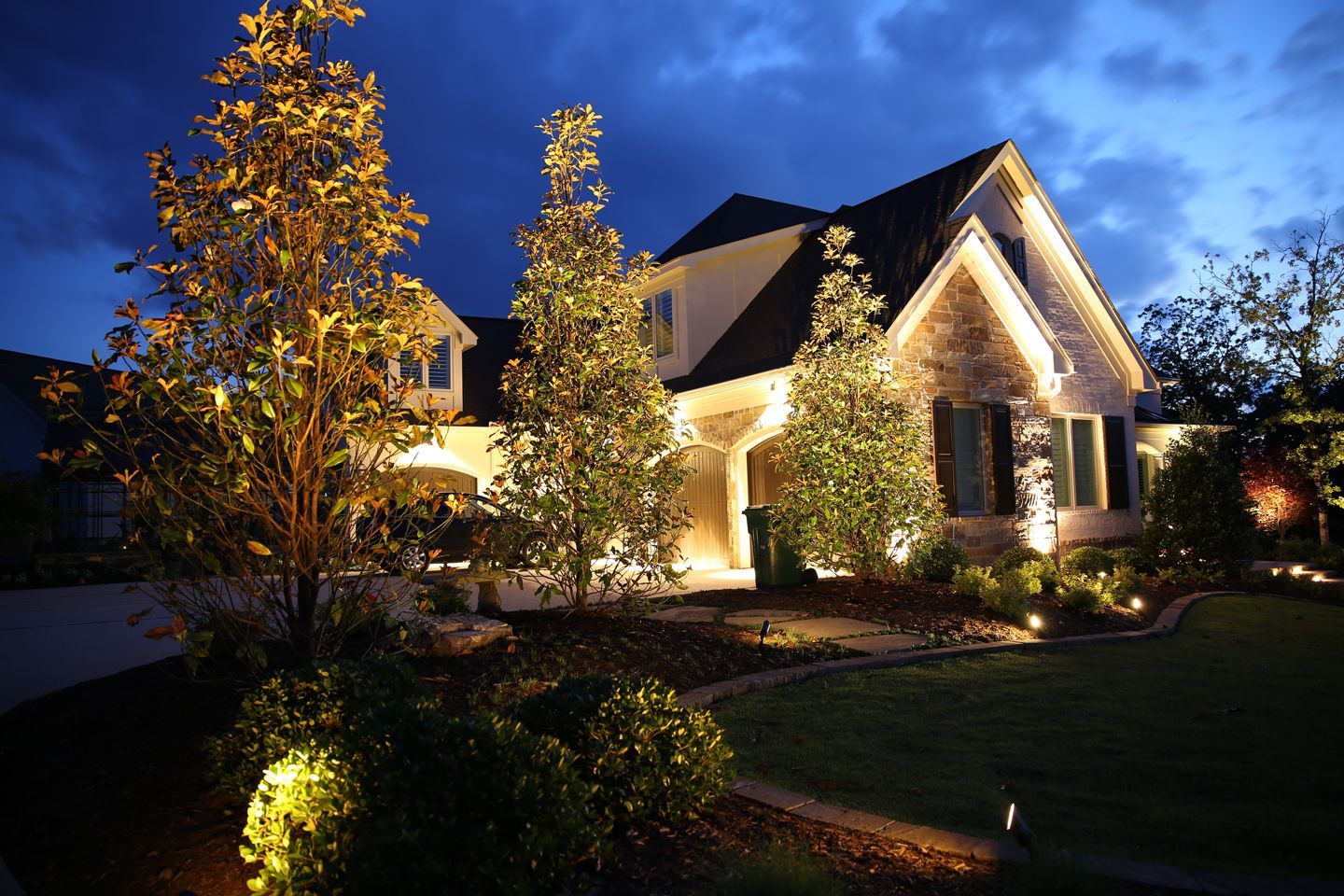 Well-lit suburban house at dusk with warm exterior lights and landscaped front yard