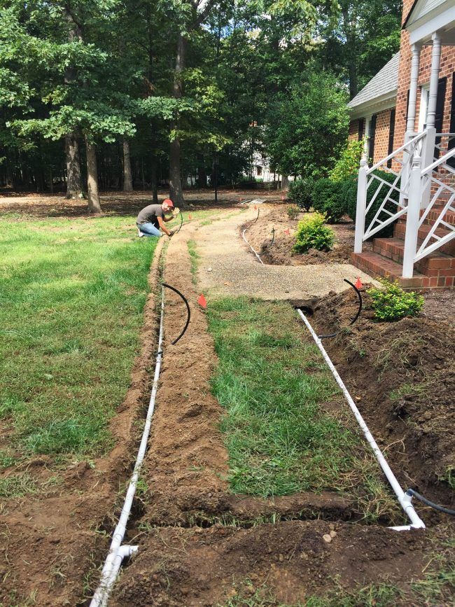 Person watering a narrow garden bed beside a house with white edging and a brick path beyond.