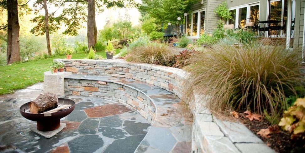 Curved stone patio with a small fire bowl, landscaped plants, and a house beside a garden path