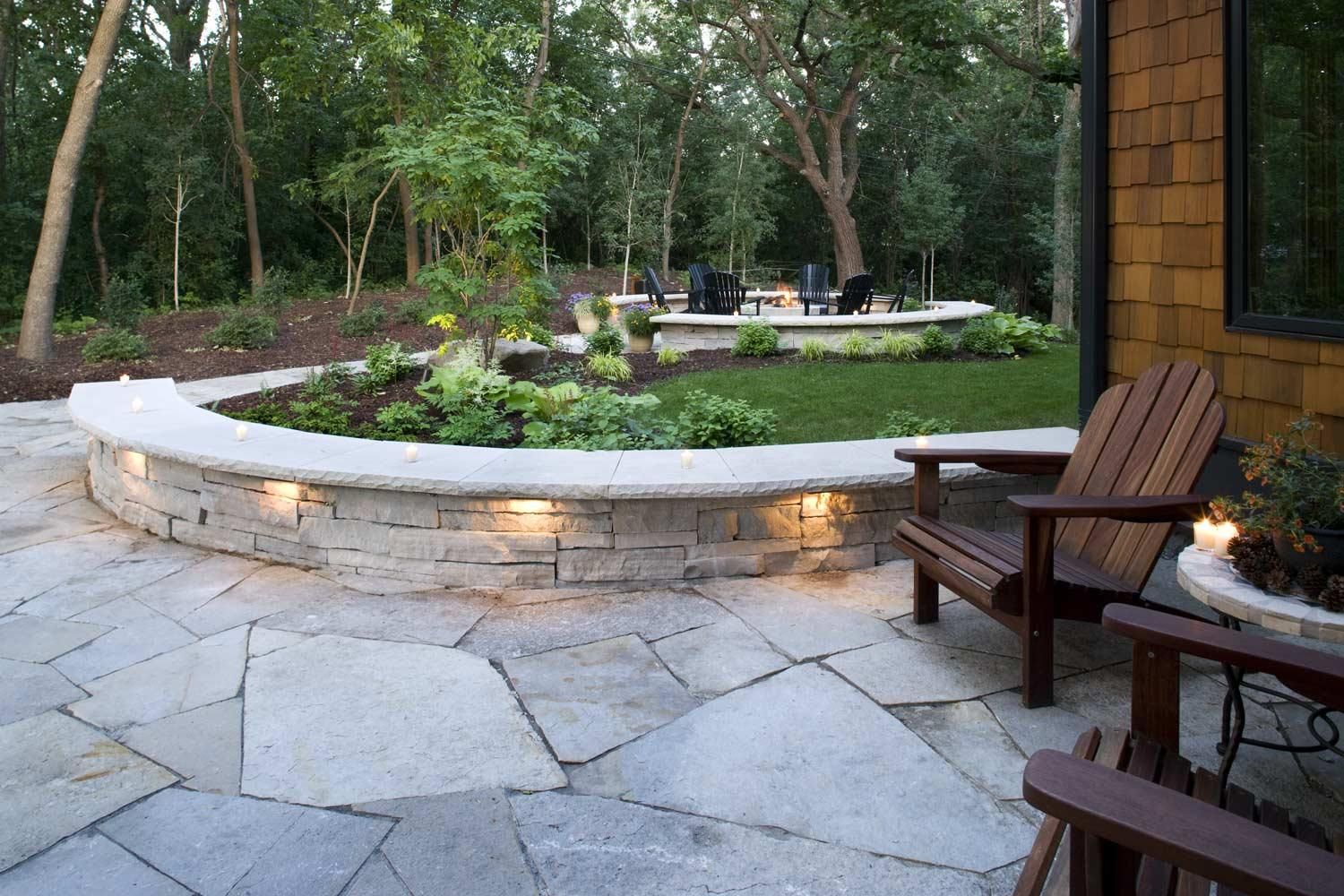 Stone patio and curved illuminated planter in a wooded garden beside wooden chairs and a house.