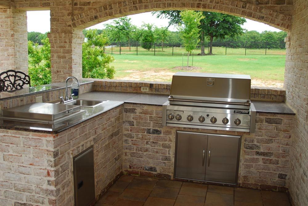 Brick outdoor kitchen with stainless steel grill and sink under a stone-covered patio overlooking a green field