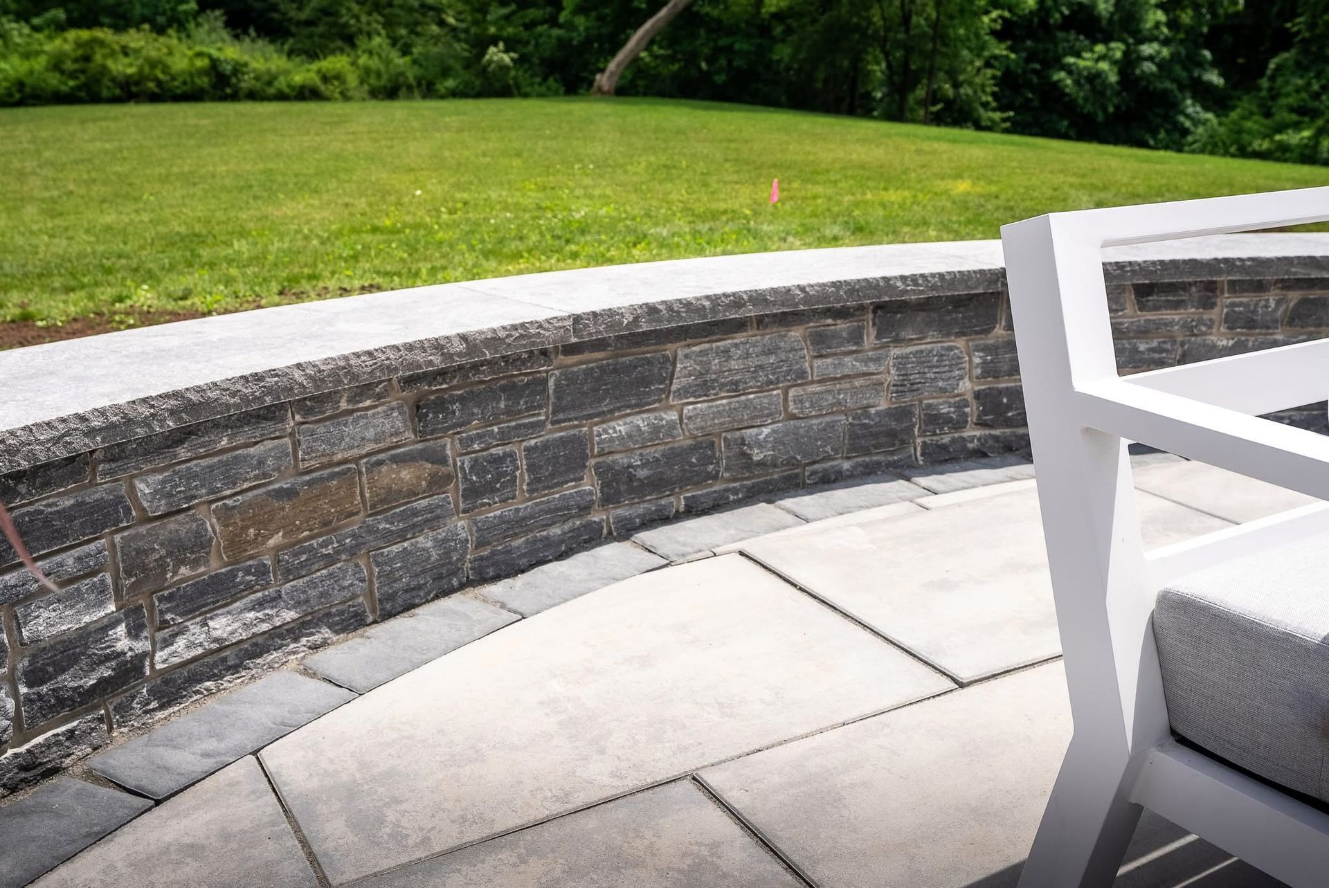 Curved stone retaining wall beside a paved patio and green lawn, with a white chair in the foreground