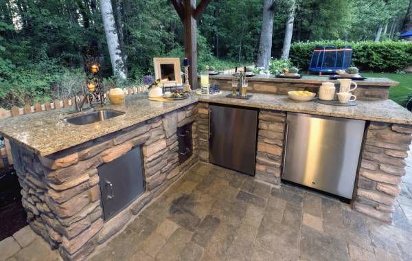 Outdoor stone kitchen with granite counters, sink, mini fridge, and grill in a wooded backyard