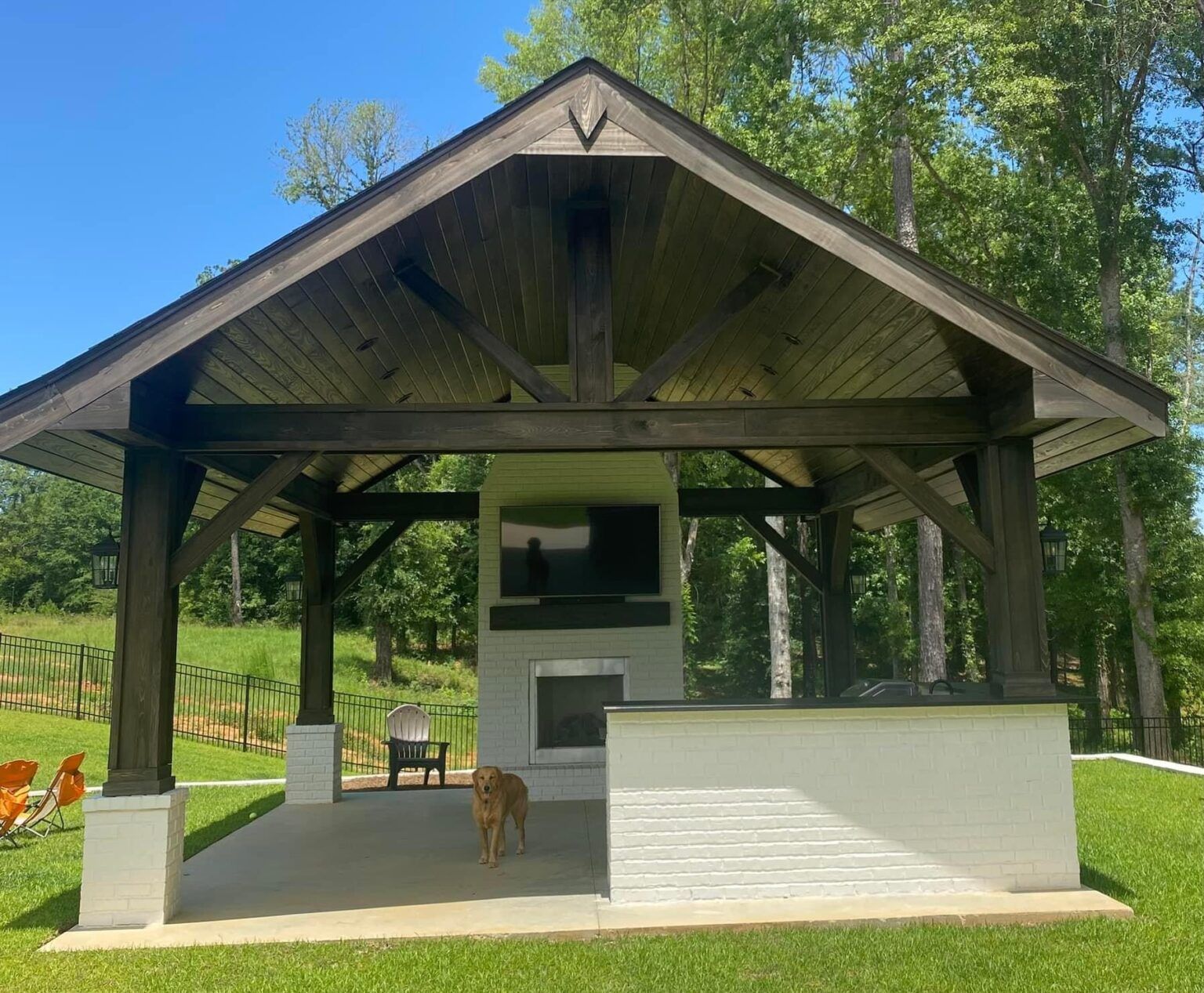 Open-air pavilion with a fireplace, white bar counter, and chairs on a grassy lawn by trees