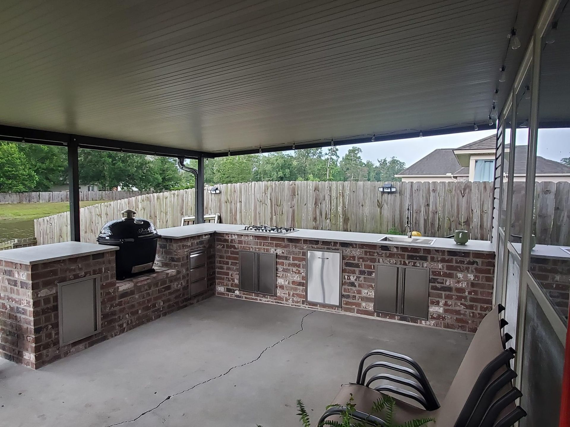 Covered brick outdoor kitchen under a patio with grill, countertop, and chairs