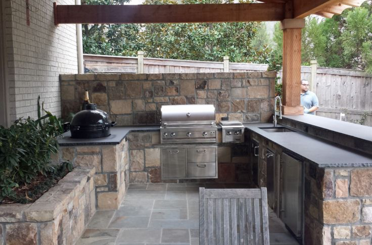 Outdoor stone patio kitchen with built-in grill, sink, counter, and bar seating under a wooden roof