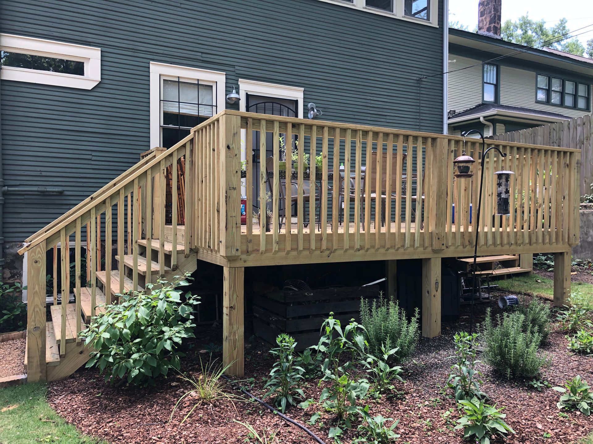 Wooden backyard deck with railings and stairs beside a house, surrounded by shrubs and mulch
