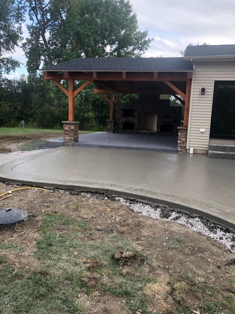 Concrete patio beside a covered carport with stone columns and a grassy yard.