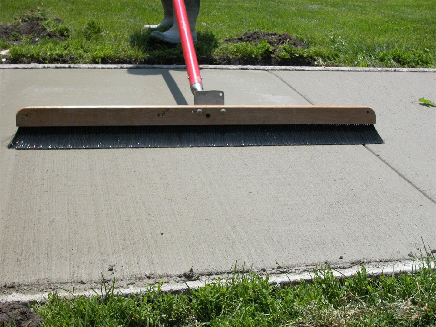 Red-handled broom smoothing wet concrete on a sidewalk near grass