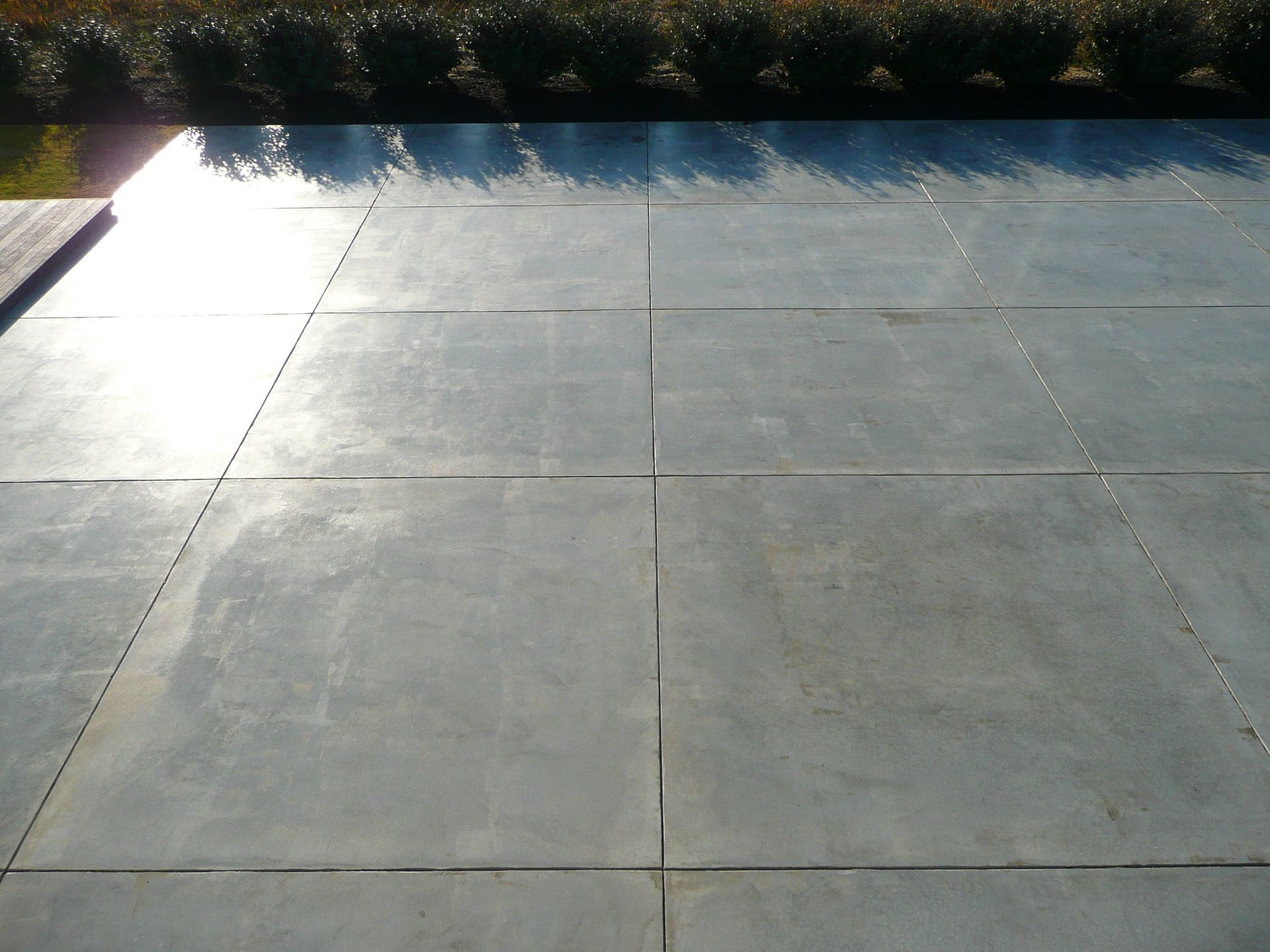 Empty gray concrete patio with a bright sunlit reflection and shrubs along the back edge