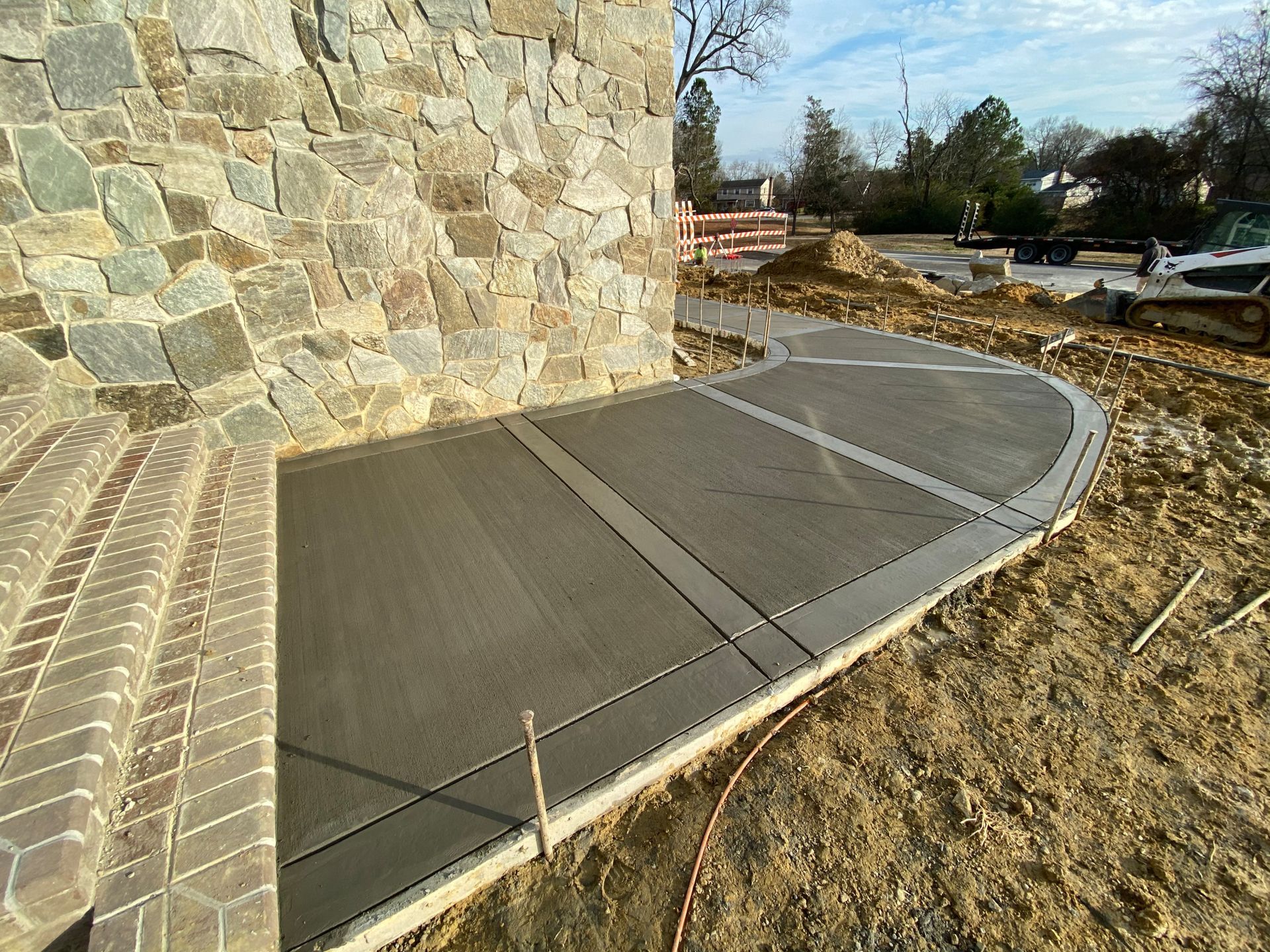 Concrete sidewalk beside a stone wall, with a curved curb and dirt construction area nearby