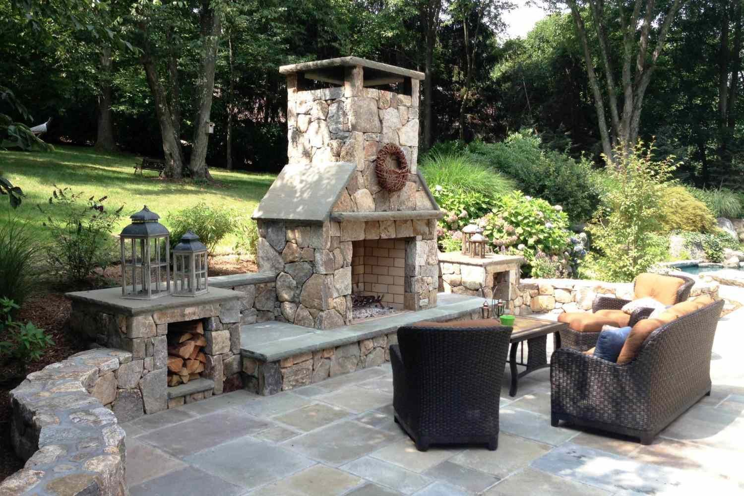 Stone outdoor patio with fireplace, wicker chairs, and lush garden greenery