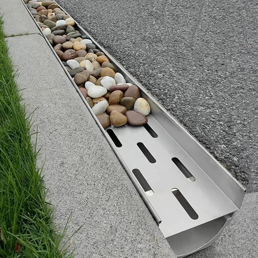 Metal drainage channel filled with smooth river rocks along a curb beside pavement and grass