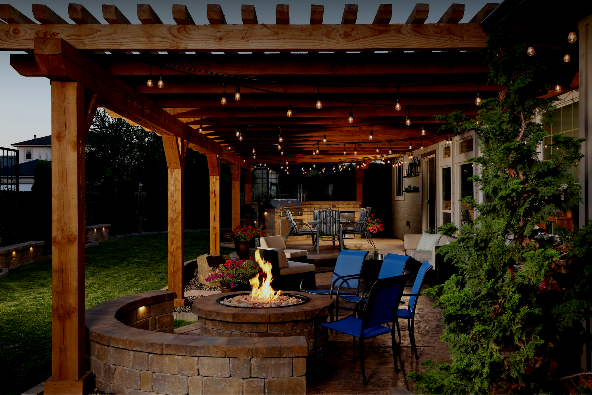 Covered patio with a stone fire pit, blue chairs, string lights, and greenery at dusk