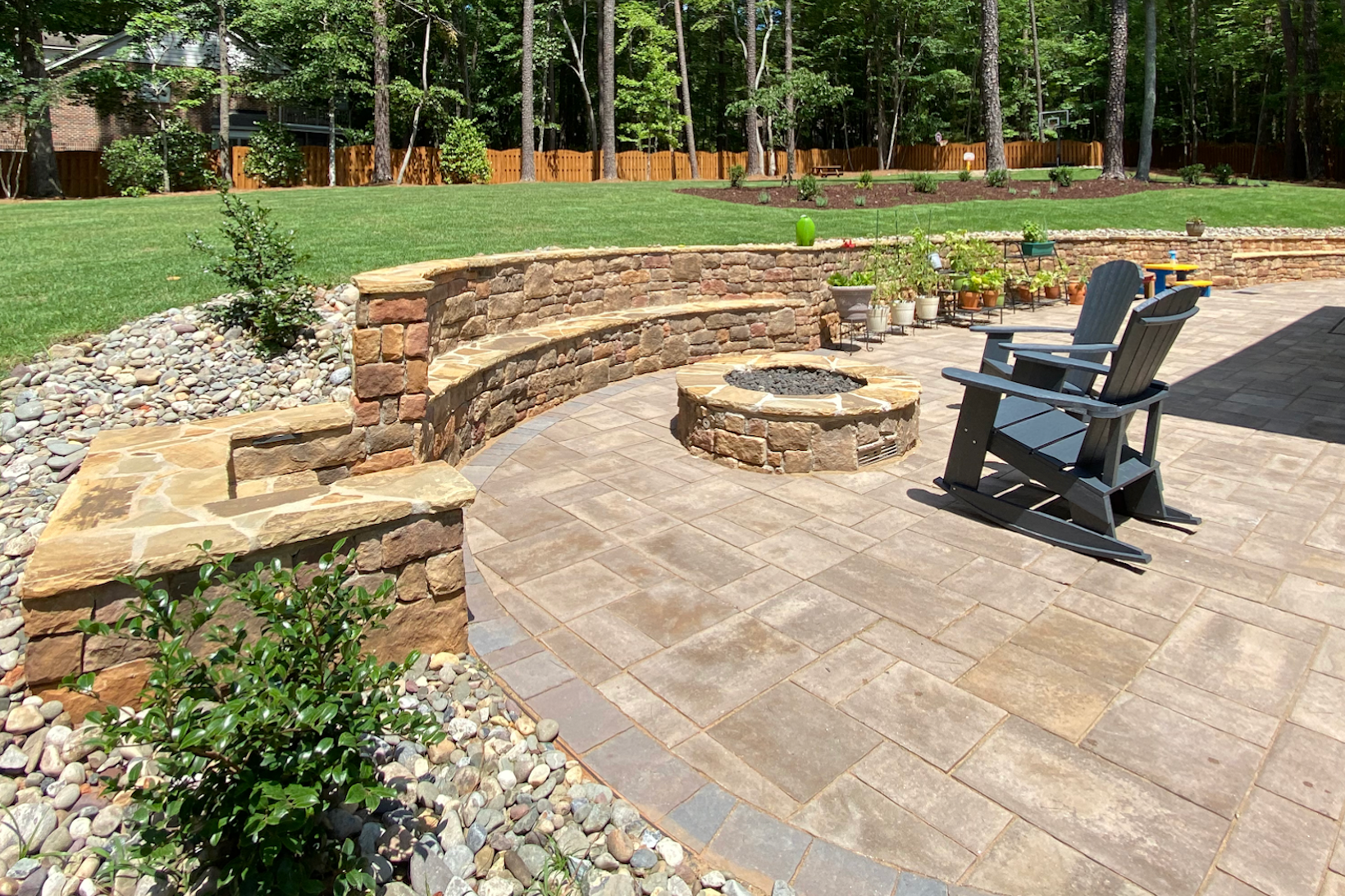 Stone patio with curved retaining wall, fire pit, and two black chairs in a landscaped backyard