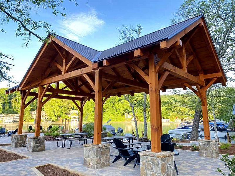 Wooden pavilion with stone columns and picnic tables in a park, with trees and parked cars in the background