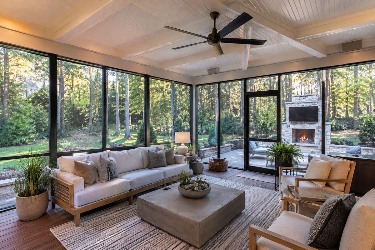 Bright sunroom with gray sofas, a coffee table, wicker chairs, and floor-to-ceiling windows overlooking trees