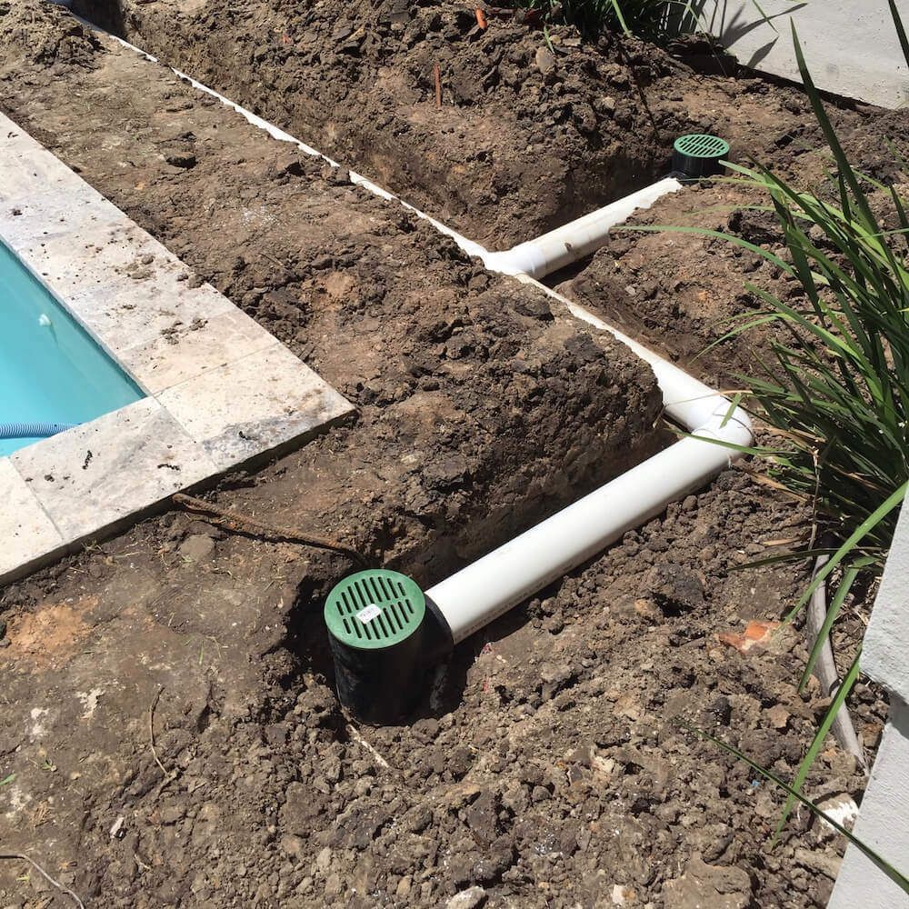 PVC pool drainage pipes in a dirt trench beside a pool, with green-capped vents.
