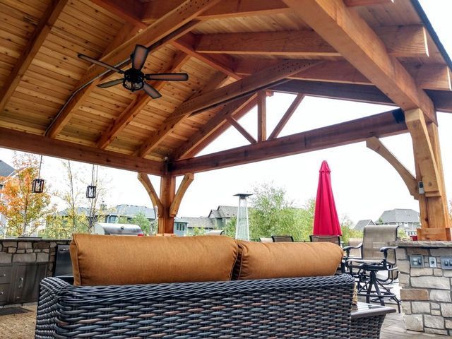 Covered outdoor patio with a wicker sofa, tan cushions, red umbrella, and ceiling fan under a wooden roof