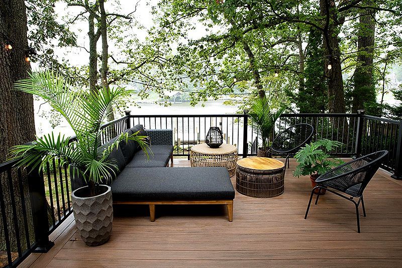 Wooden deck with black lounge chair, wicker fire pit, and potted plants overlooking a lake