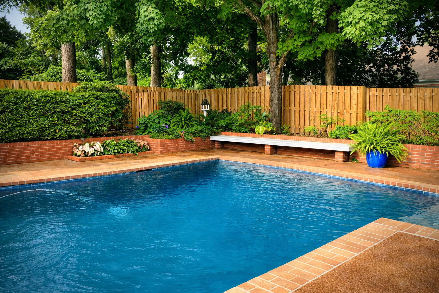 Backyard swimming pool with blue water, brick deck, wooden fence, and lush green trees and plants