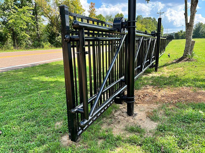 Black metal farm gate beside a roadside, standing open in a grassy area under trees