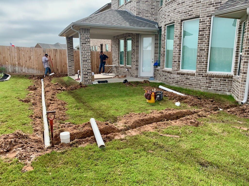 Brick house backyard under construction with muddy trenches, pipes, and two workers near the patio.