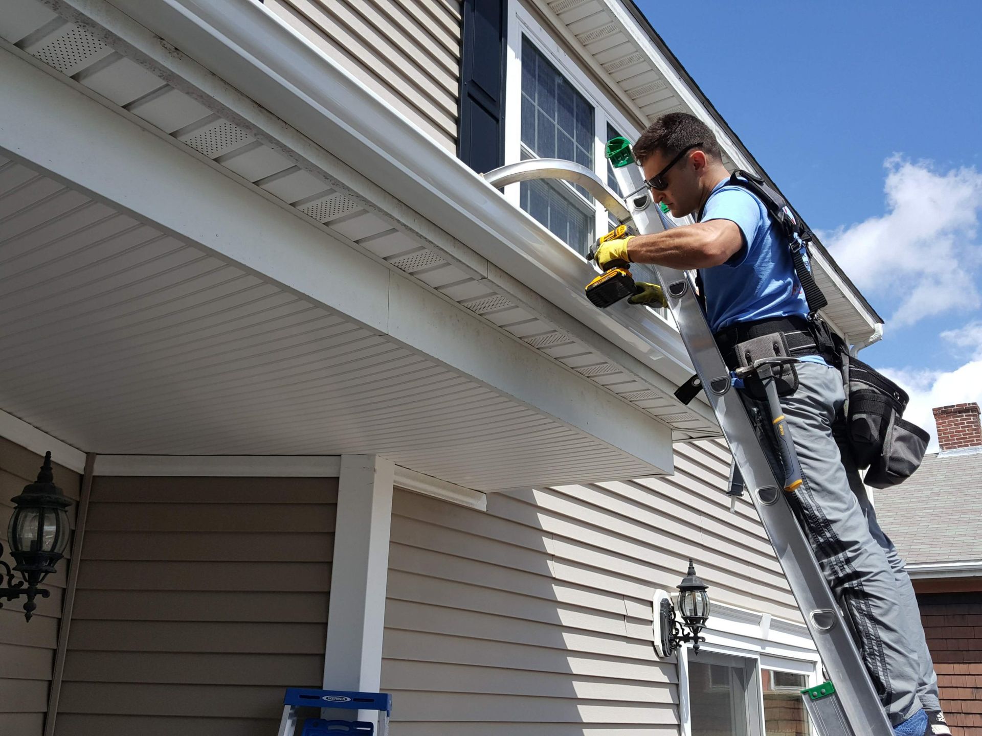 Worker using a power tool on a house roofline, standing on a ladder under a sunny blue sky