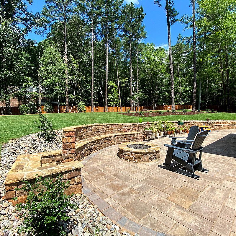 Backyard patio with stone fire pit, curved seating wall, and Adirondack chair beside a wooded lawn
