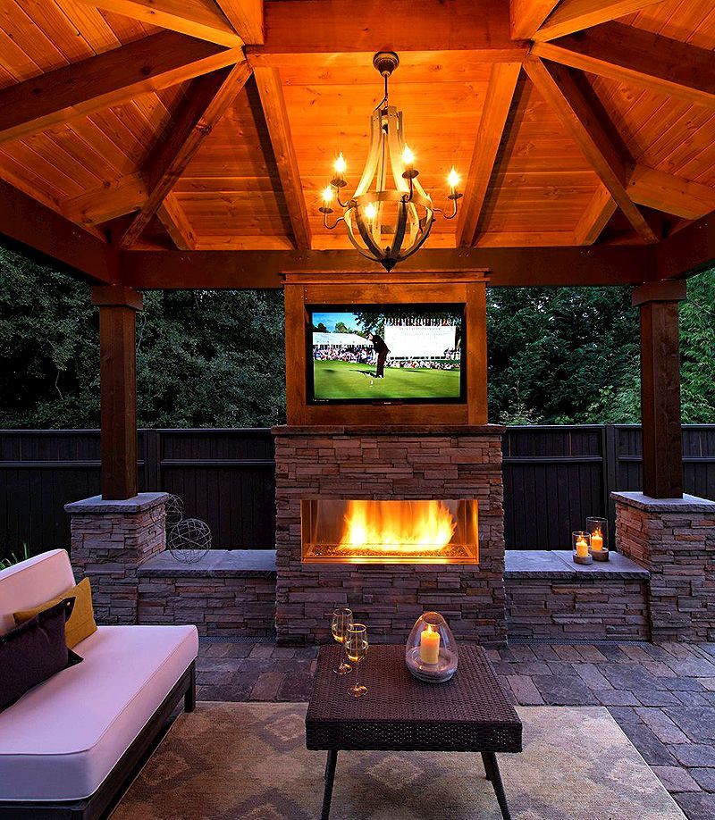 Cozy covered patio with stone fireplace, mounted TV, chandelier, and cushioned seating at dusk