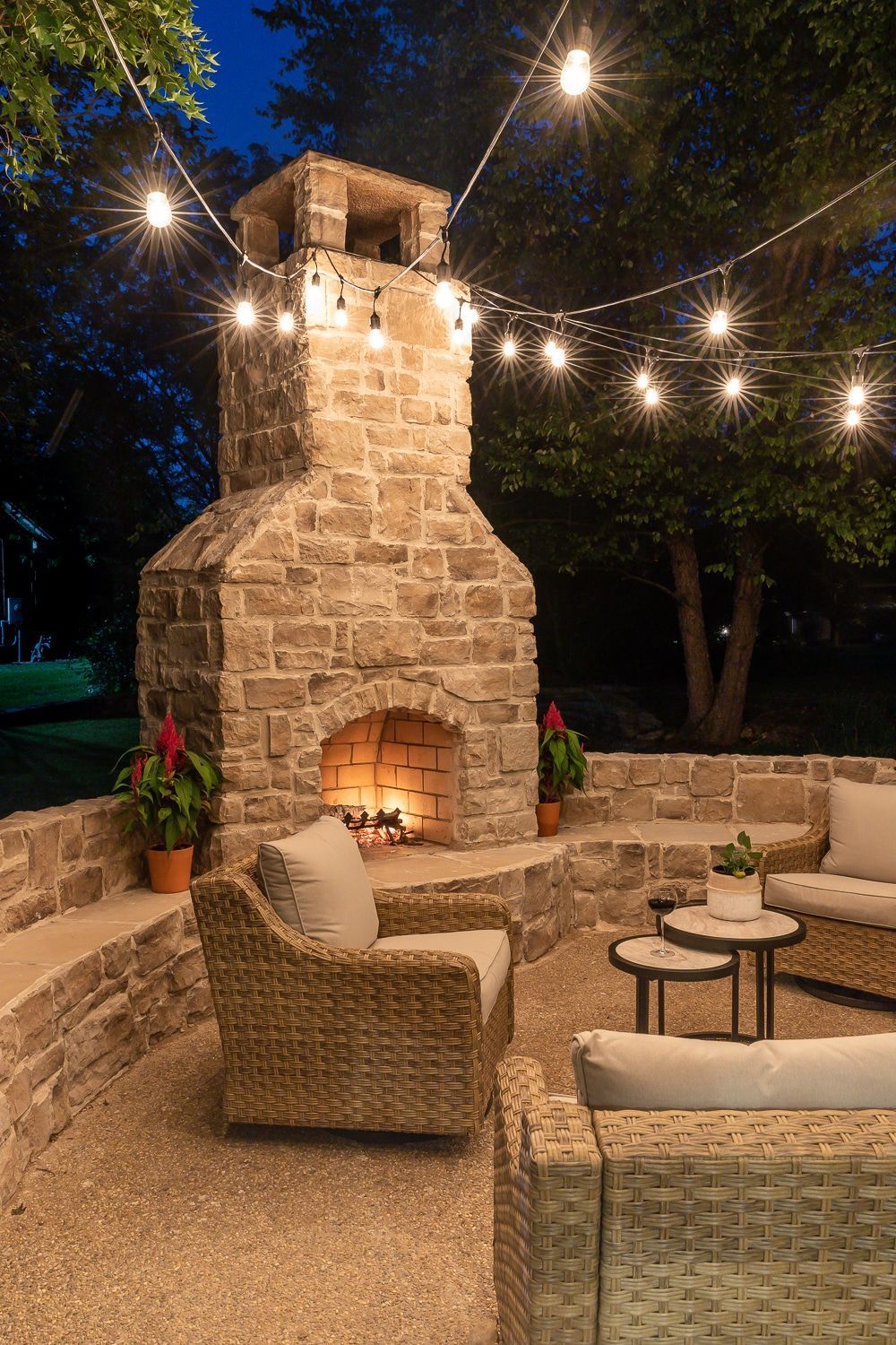 Outdoor stone fireplace on a patio at night, surrounded by wicker seating and string lights