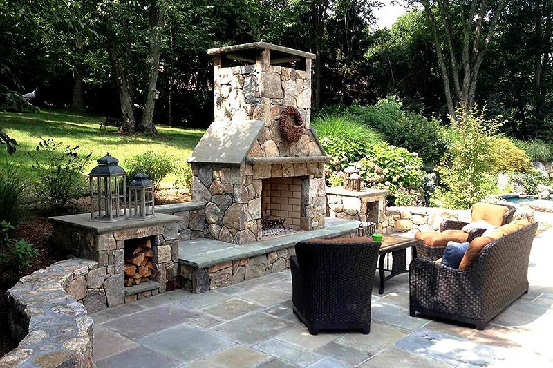 Stone outdoor fireplace with wicker chairs and a black fire pit on a patio surrounded by greenery
