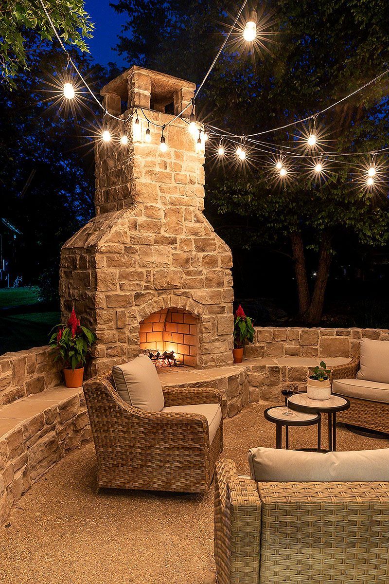 Stone outdoor patio with a lit fireplace, wicker chairs, and string lights at dusk