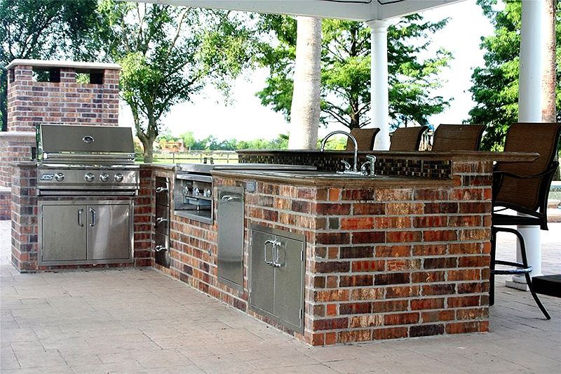 Outdoor brick-built grill and bar under a pavilion with stainless steel appliances and chairs
