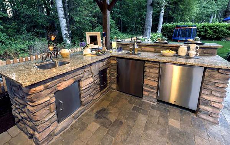 Outdoor stone kitchen with granite counters, sink, and built-in grill in a wooded backyard