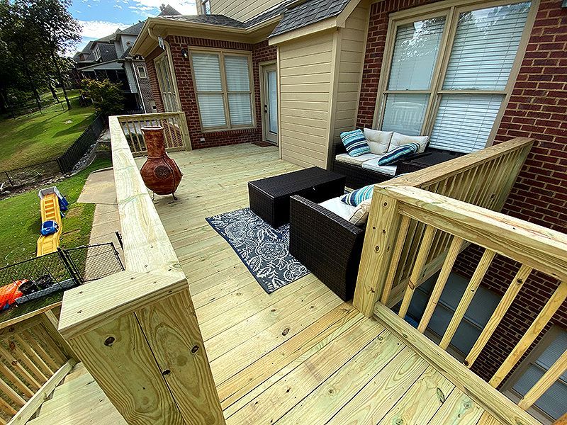 Wooden backyard deck with black patio furniture, rug, potted planter, and brick house exterior and windows