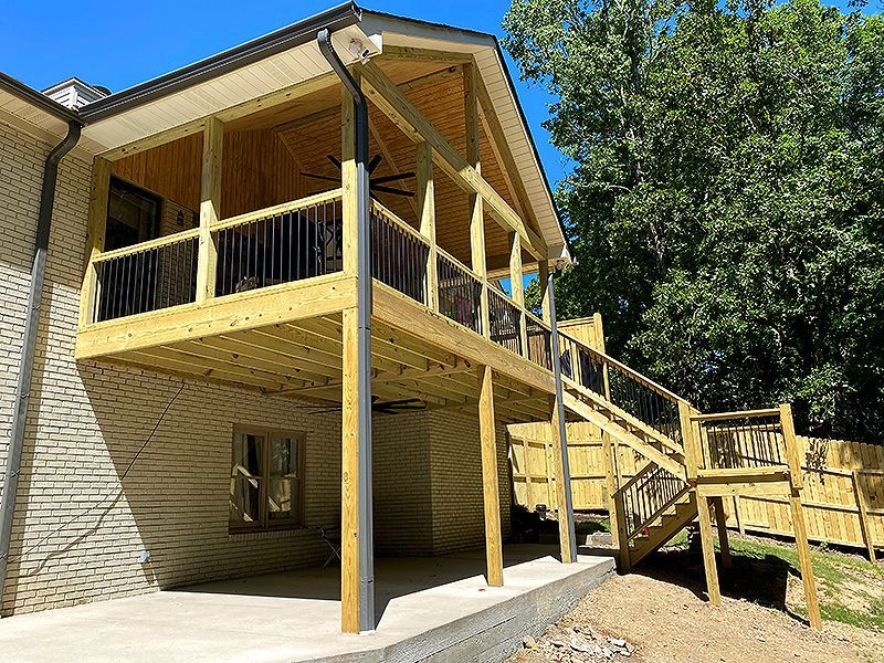 Raised wooden deck and staircase attached to a beige house beside trees under a blue sky