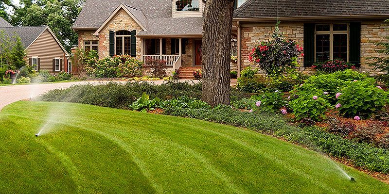 Sprinkler watering a lush green lawn in front of a brick house with colorful garden beds