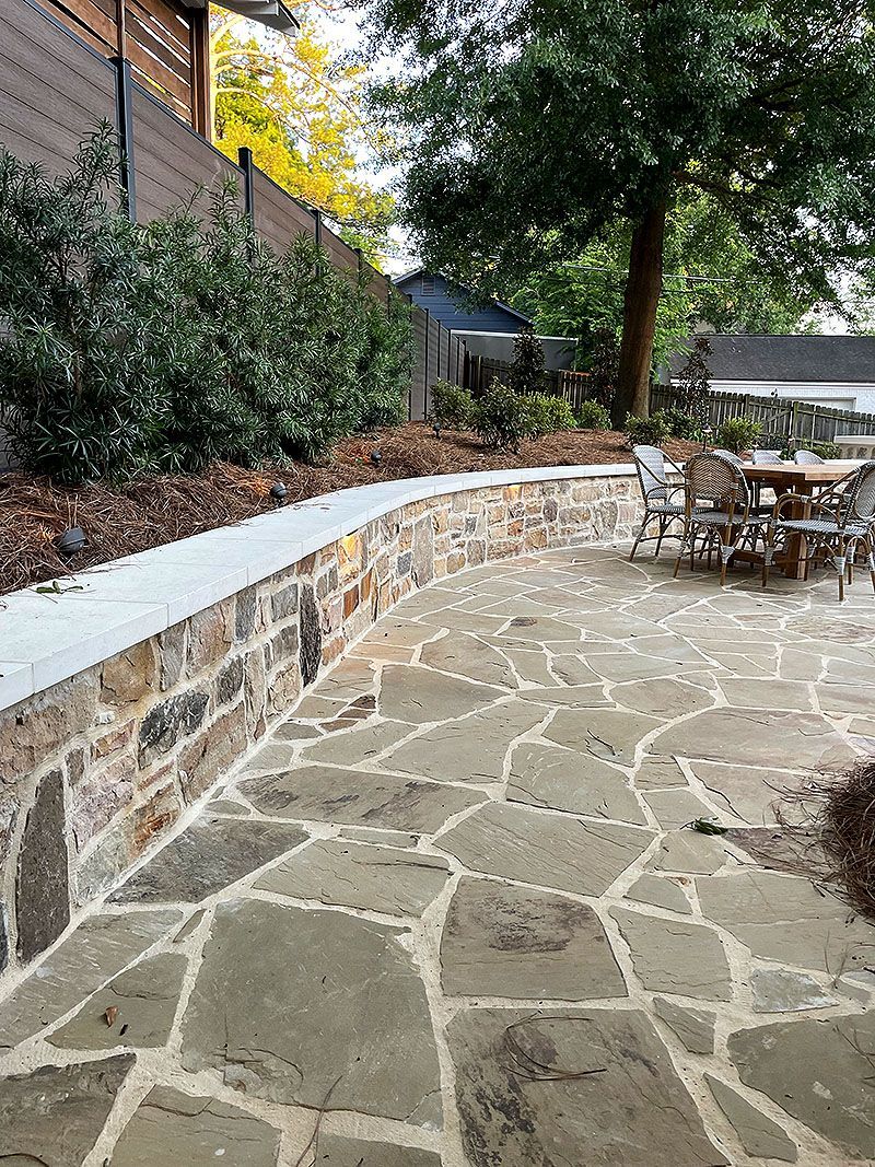 Stone patio with curved retaining wall, outdoor table and chairs, trees, and landscaped garden