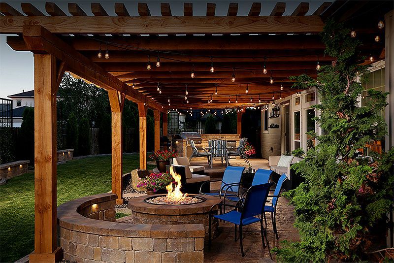 Covered patio with wooden pergola, string lights, and a stone fire pit surrounded by blue chairs at dusk