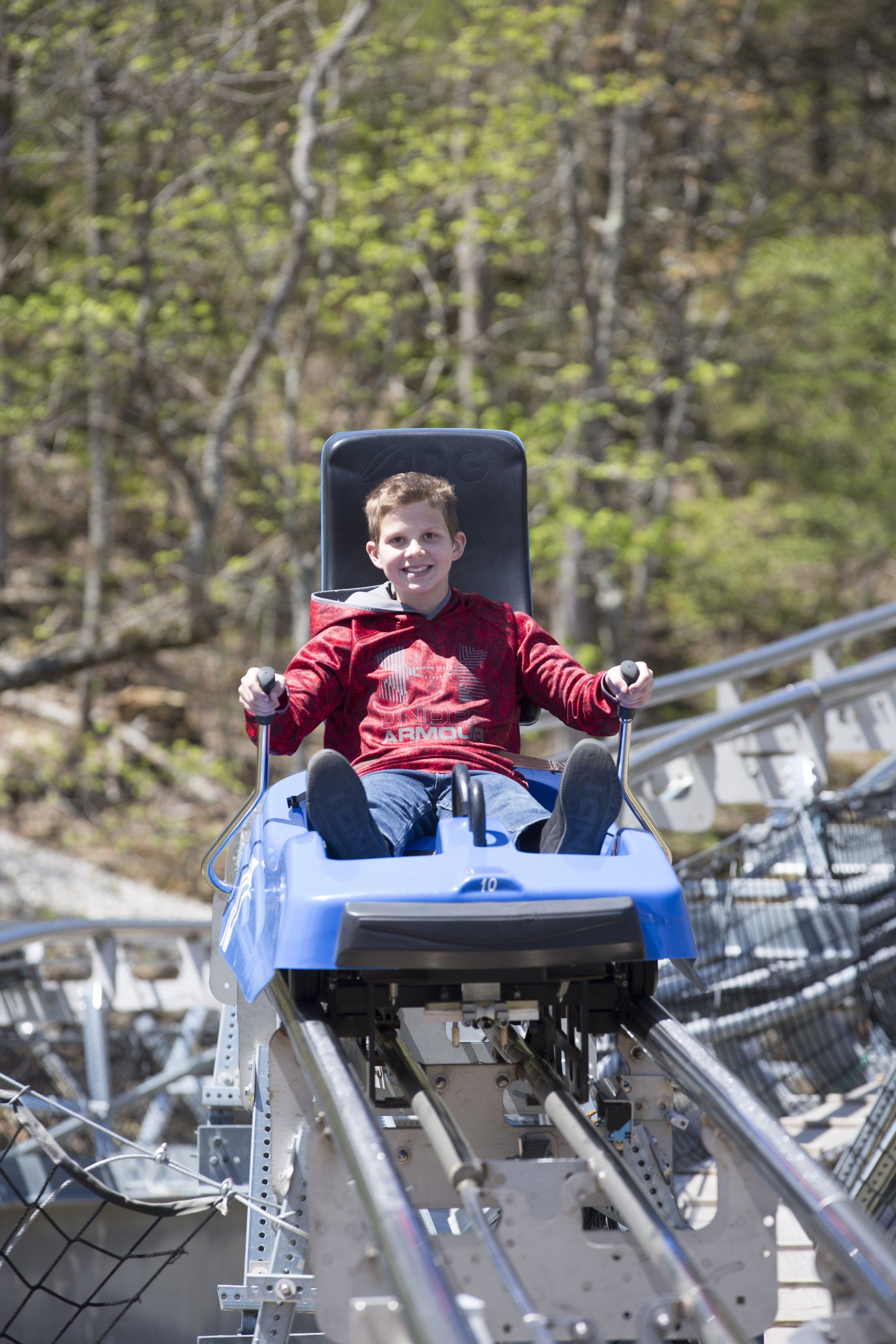 Young boy riding a blue alpine coaster cart through the woods, smiling with excitement