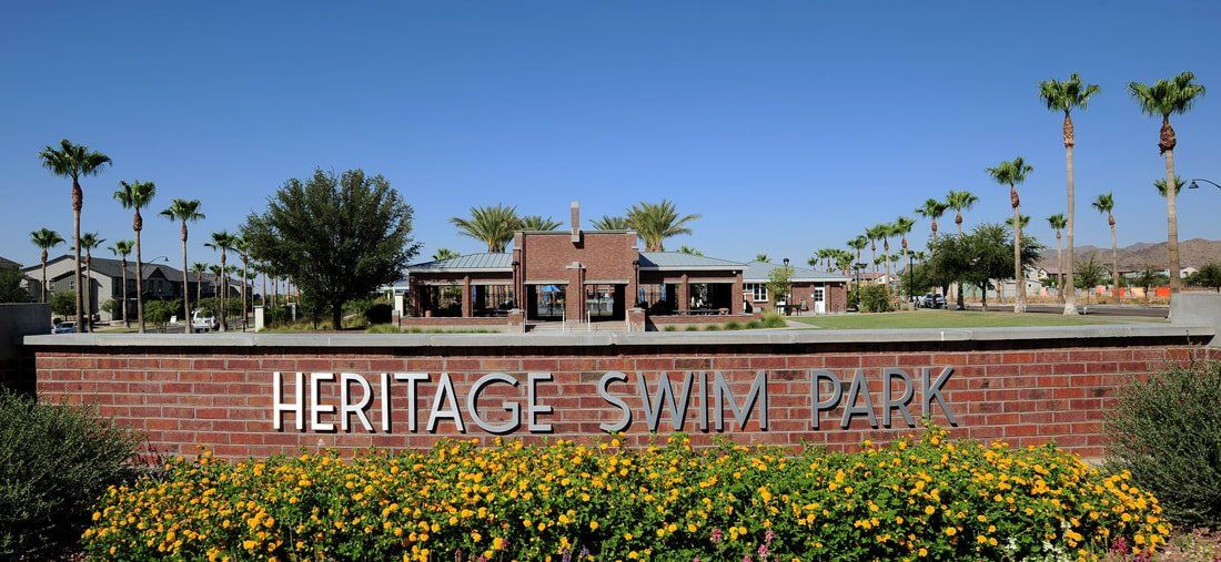 A sign for heritage swim park is surrounded by flowers and palm trees.